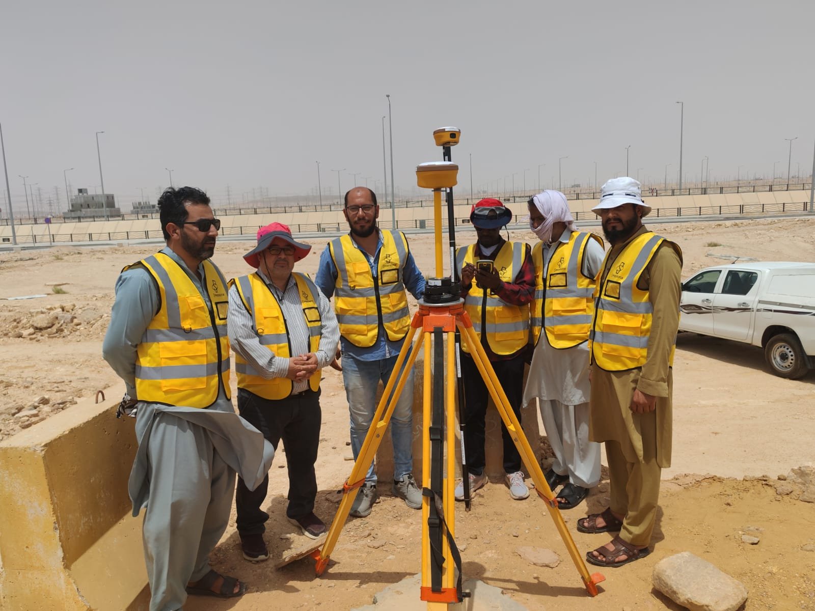 ستة أعضاء من فريق مساحي يرتدون سترات السلامة بجانب جهاز GNSS في موقع ميداني
