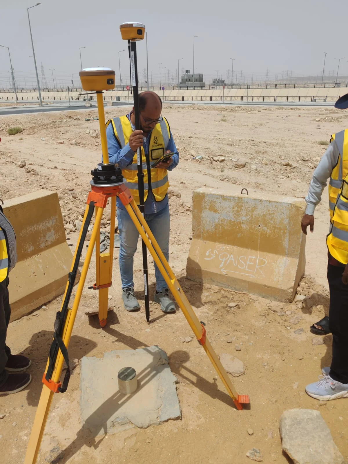 فني مساحي يرتدي سترة سلامة صفراء يراجع جهازًا محمولًا بجوار معدات GNSS