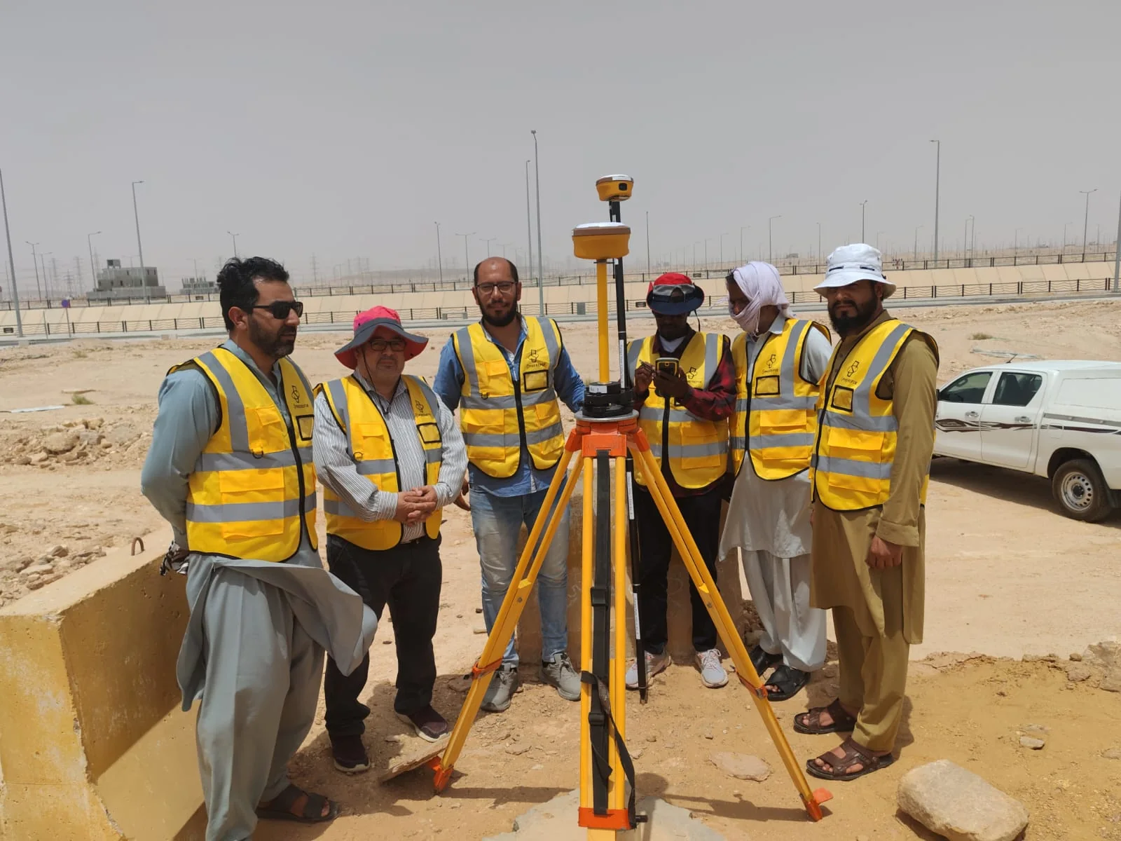 ستة أعضاء من فريق مساحي يرتدون سترات السلامة بجانب جهاز GNSS في موقع ميداني